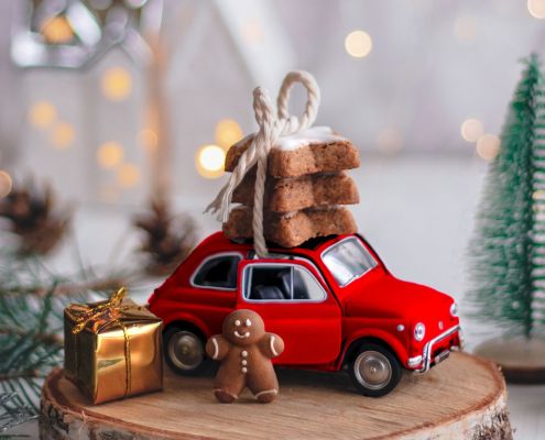 Auf einem Holzstamm ein kleines rotes Auto mit Keksen auf dem Dach, ein Gingerbredman und ein kleines Geschenk in Goldpapier